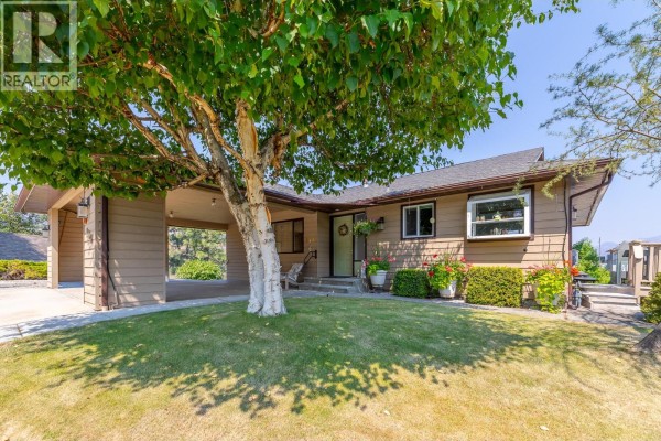 Front exterior of a house with beige siding, a covered carport, and a large tree. - 129 Sumac Ridge Drive, Summerland
