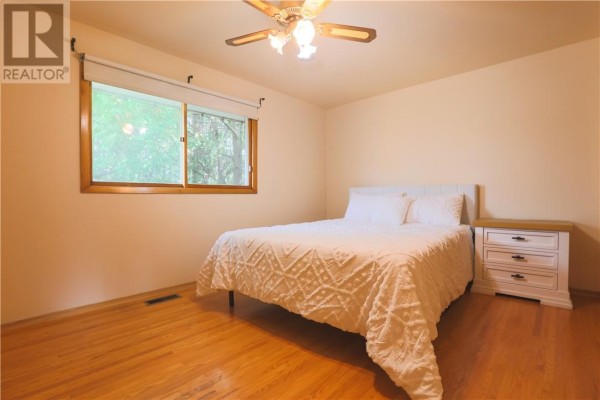 Bedroom with white bed, wooden nightstand, hardwood floors, and large window - 1295 Gemmell Street Sudbury
