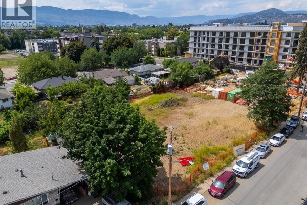 View of the undeveloped lot from a low angle, bordered by trees, homes, and a street with cars. - 1299-1329 Pridham Avenue, Kelowna