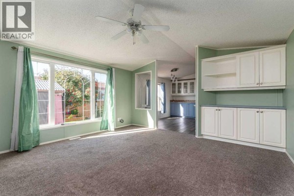 Living room with grey carpet, pale green walls, built-in cabinets, and large windows overlooking the yard. - 13, 5302 56b Street, Lloydminster