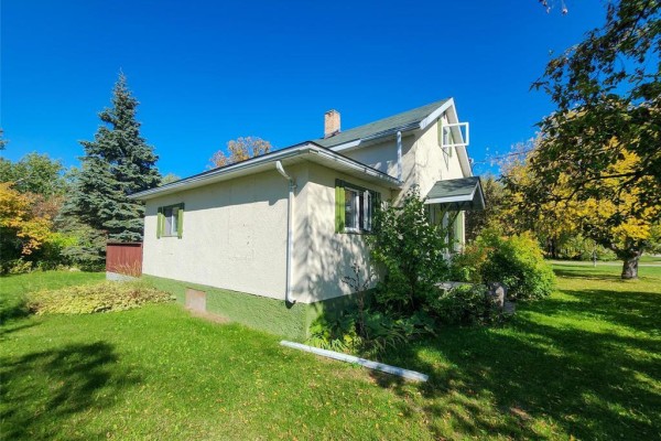 Side view of the cream-colored house showing a portion of the roof and surrounding greenery. - 13 1st Street N, Eriksdale