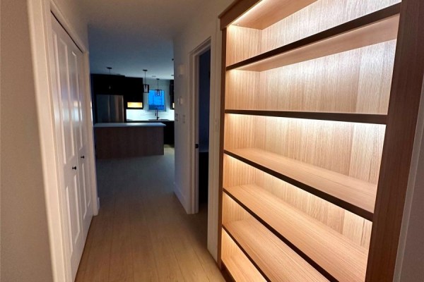 Hallway with doors to closet and kitchen, leading to a bright wood bookshelf with interior lighting. - 13 Blandford Street, Gander