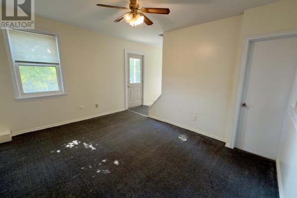 Living room with dark grey carpet, white walls, a ceiling fan, and a door to stairs. - 13 Chester Avenue, Kentville