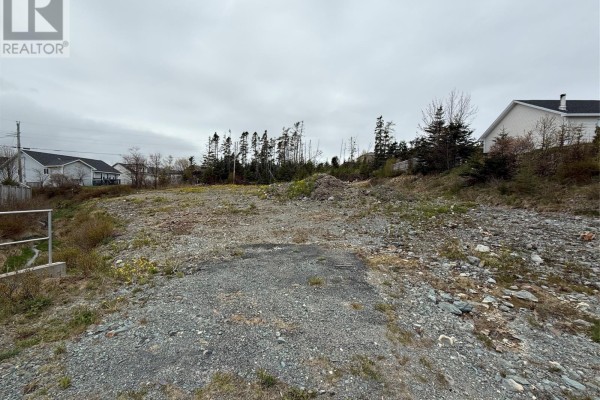 The vacant land features a gravel driveway area with surrounding rock and brush under a cloudy sky. - 13 Shea Place, Bay Roberts