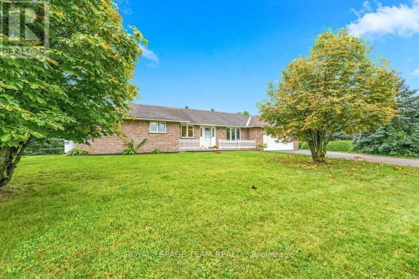 Wide shot of a brick bungalow from the front lawn, surrounded by green grass and trees. - 13 Springfield Drive, Whitewater Region