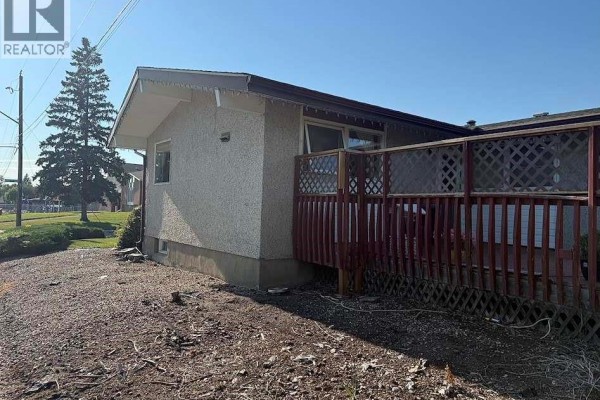 Rear side view of a bungalow with a wooden deck and fencing, adjacent to a paved area. - 1302 112th Street, North Battleford