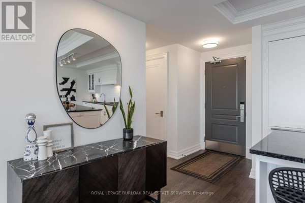 Apartment entryway with a dark wood cabinet, abstract mirror, and gray front door. Kitchen visible in background. - 1303 - 2060 Lakeshore Road, Burlington