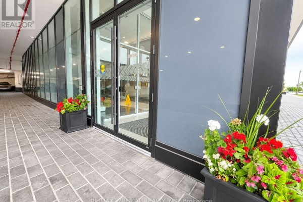 Building entrance with glass doors, gray stone paving, and planters with red and white flowers. - 1303 - 2481 Taunton Road, Oakville