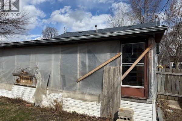 Close-up side exterior view of a house with white siding covered by clear plastic sheeting, wooden braces, and a brown door with a glass pane. - 1303 Central, Windsor