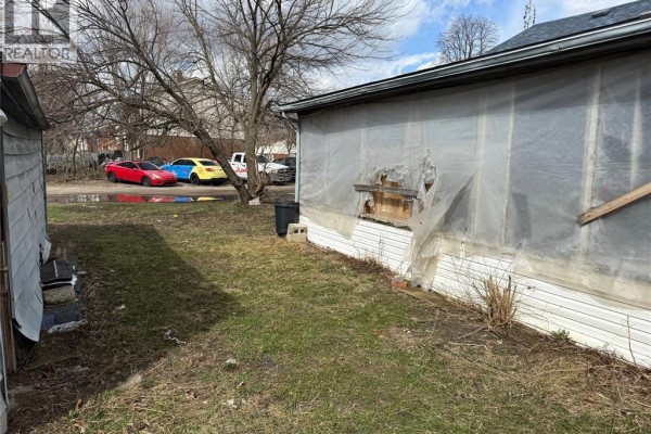 Side of house with damaged siding and plastic sheeting, grassy yard with cars - 1303 Central Windsor