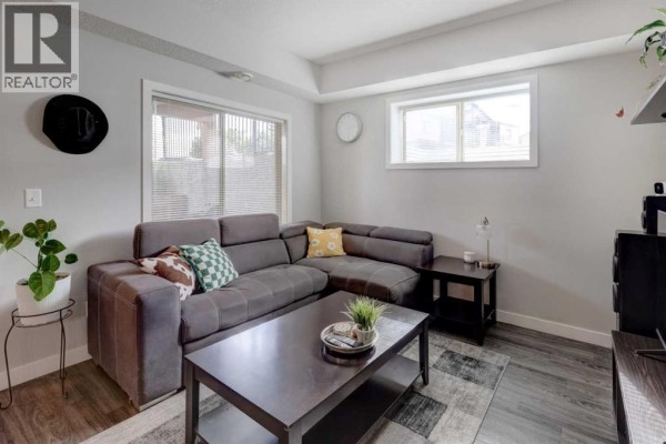Living room with a grey sectional sofa, dark coffee table, and wood-look flooring. - 1304, 2461 Baysprings Link Sw, Airdrie