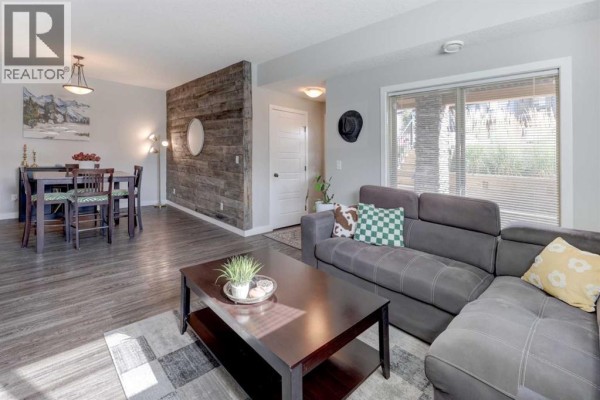 Living and dining area with a grey sectional, wood-accent wall, and dining table. - 1304, 2461 Baysprings Link Sw, Airdrie
