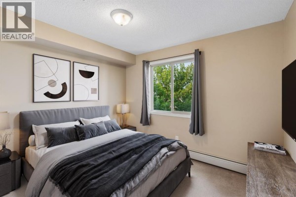 Bedroom with a king-sized bed, grey bedding, and a window with green trees outside. - 1305, 8 Bridlecrest Drive Sw, Calgary