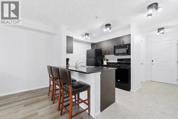 Living room with black sofa, white coffee table, rug, and view into the open kitchen area. - 1308, 625 Glenbow Drive, Cochrane