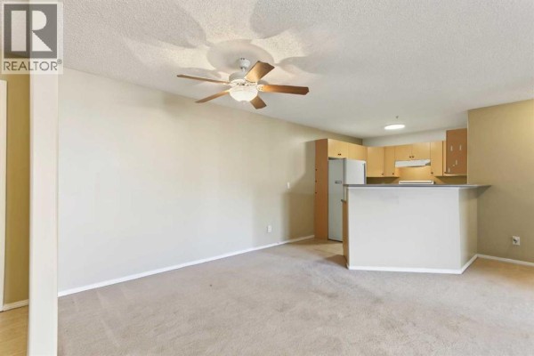 Living room with beige carpet, ceiling fan, and an open-concept kitchen with light wood cabinets. - 1310, 16320 24 Street Sw, Calgary