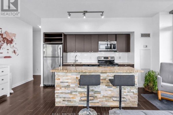 Kitchen with dark wood cabinets, stainless steel appliances, granite countertops, and a stone-clad island with bar stools. - 1310 - 105 The Queensway, Toronto