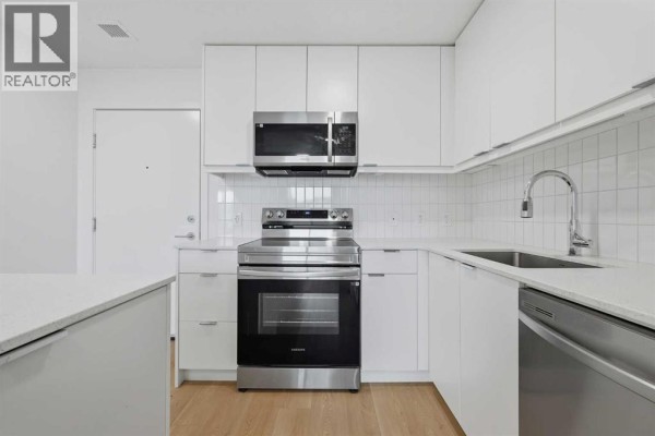 Corner view of a modern white kitchen featuring stainless steel appliances, white cabinetry, and a light wood floor. - 1313, 8500 19 Avenue Se, Calgary