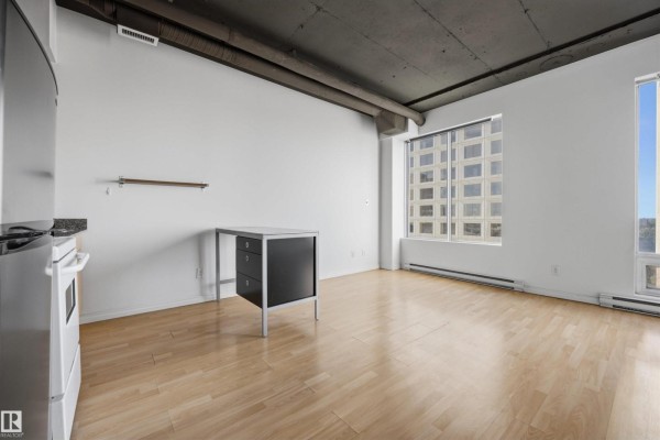 Studio apartment with light wood floors, white walls, concrete ceiling, stainless steel appliances, and a large window facing a city building. - #1313 10024 Jasper Av Nw, Edmonton