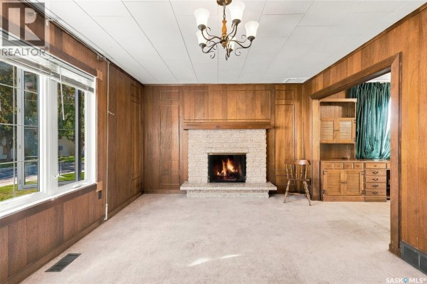 Living room with a fireplace, wood paneling, a chandelier, and large windows. - 1315 2nd Avenue N, Saskatoon