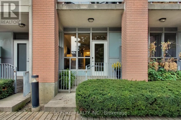 Exterior view of the apartment entrance with brick facade, glass doors, and lush green hedges. - 132 - 1005 King Street W, Toronto