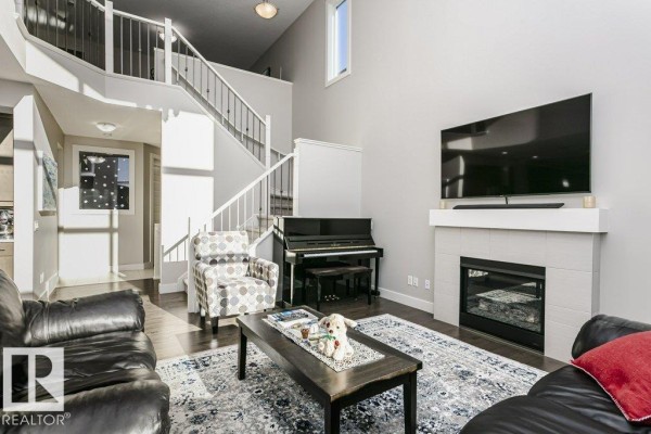 Living room with a black upright piano, a gas fireplace, a patterned armchair, and a staircase leading to the upper floor. - 1327 158 St Sw, Edmonton