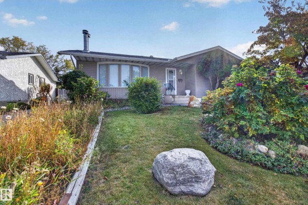 Front exterior of a beige bungalow with a sloped lawn, mature tree, and flower beds. - 13315 30 St Nw, Edmonton