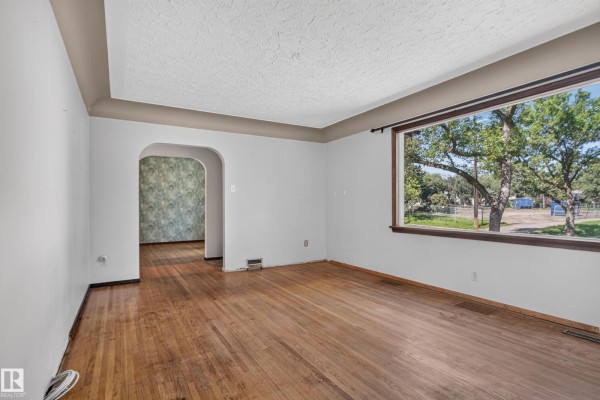 Living room with hardwood floors, white walls, an arched doorway, and a large window showing greenery. - 13351 109a Av Nw, Edmonton