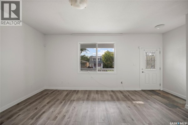 Living room interior with white walls, grey wood-look flooring, a large window, and a door. - 1337 Athol Street, Regina