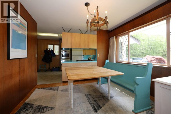Interior dining area with wood-paneled walls, a wooden table, a turquoise built-in bench, and a large window with garden views. - 1346 Russell Road, Gibsons
