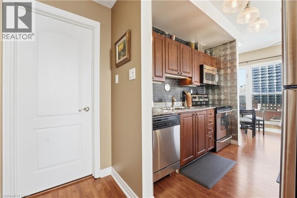 Apartment interior with hallway door, kitchen with wood cabinets, stainless steel appliances, and dining area view. - 135 James Street S Unit# 802, Hamilton