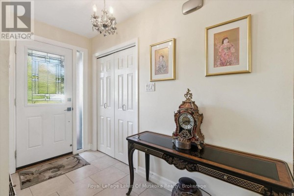 The home's foyer features beige tile flooring, a white door with glass, two framed pictures, and a decorative clock on a table. - 135 Muskoka Beach Road, Gravenhurst)