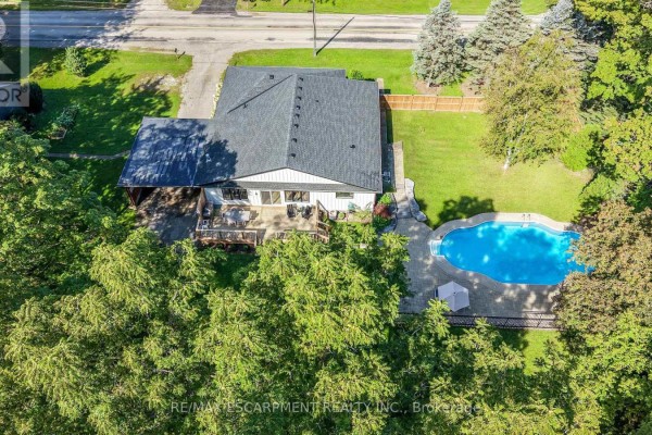 Aerial view of the backyard of a red brick bungalow featuring a wooden deck, a swimming pool, and mature trees. - 1355 Woodburn Road, Hamilton