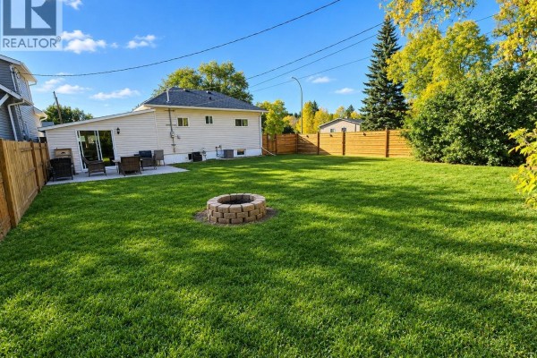 Large green backyard with a circular stone fire pit and a wooden fence. - 136 10th Street Ne, Weyburn