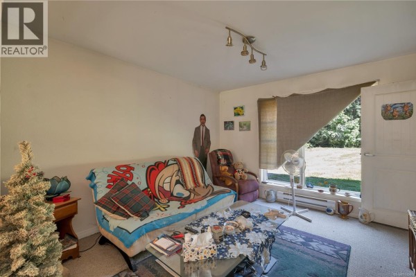 Living room with couch, coffee table, fan, and view of greenery through window. - 1376 Hooley Rd, Quadra Island