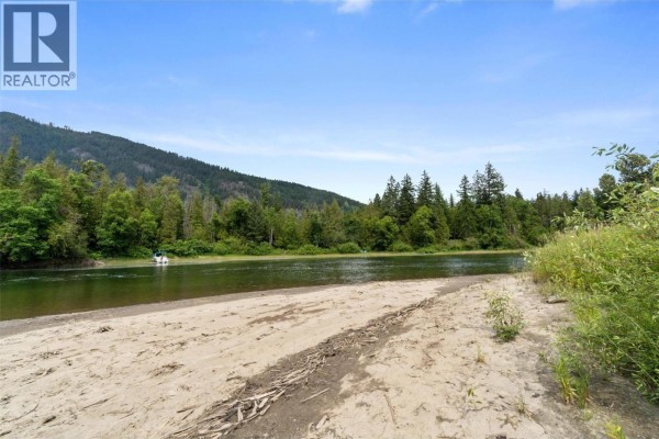 Sandy shoreline of a calm river with trees and mountains in the background. - 1383 Silver Sands Road Unit# 109, Sicamous