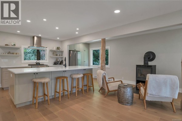 Modern open-concept kitchen and living area with a large island, stainless steel appliances, and a wood-burning stove. - 1387 Heriot Bay Rd, Quadra Island