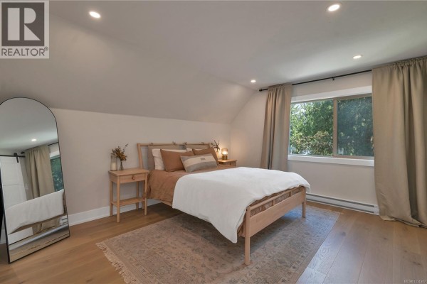 Bright bedroom with a queen-sized bed, natural wood flooring, and a large arched floor mirror. - 1387 Heriot Bay Rd, Quadra Island