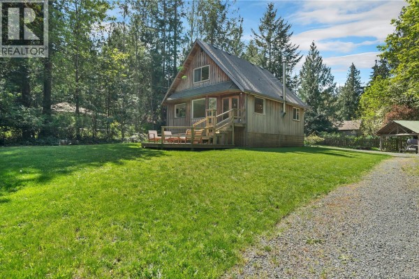Side view of the wooden cabin with a wooden deck, surrounded by a grassy lawn and tall evergreen trees. - 1387 Heriot Bay Rd, Quadra Island