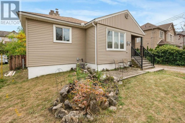 Side view of a beige vinyl-sided bungalow with a rocky, overgrown garden in the foreground. - 1388/1390 Leighland Road, Burlington
