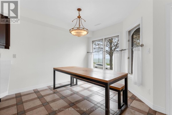 Dining area with a wooden table and bench, tiled floor, and large windows overlooking the lake. - 1389 Lakeshore Road, Burlington