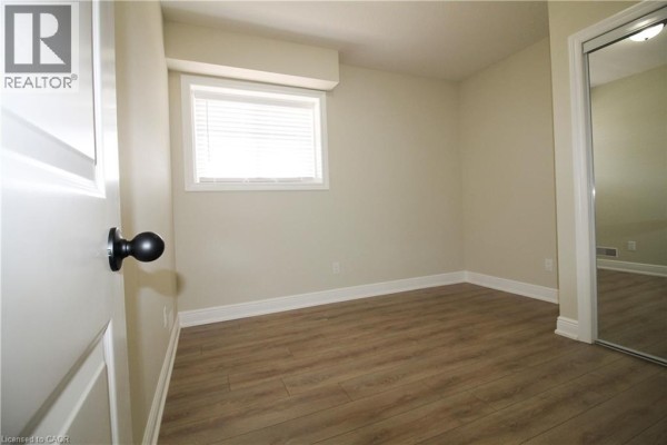Empty room with light beige walls, wood-look flooring, and a small window. - 1395 Bleams Road Unit# 8, Kitchener