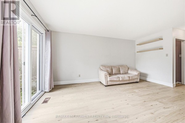 Living room with light wood laminate flooring, neutral walls, a beige sofa, and sliding glass doors with curtains. - 14 - 564 Sheraton Road, Burlington