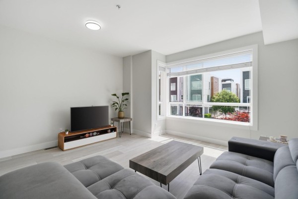 Living room with light wood floors, grey sectional sofa, coffee table, and large corner window with exterior view. - 14 19631 55a Avenue, Langley