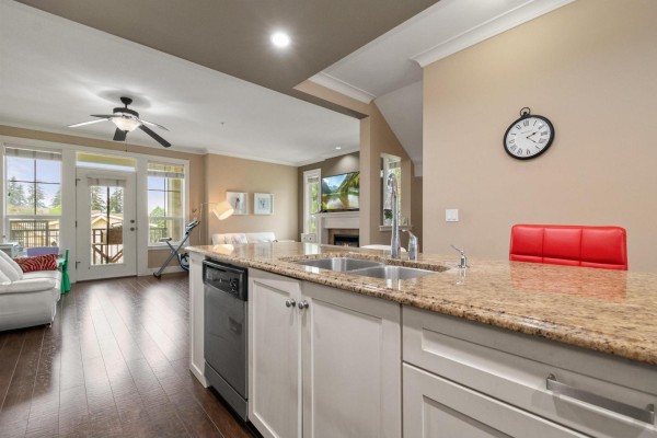 Kitchen island with sink and granite countertop, looking towards the living room with a fireplace and balcony doors. - 14 45290 Soowahlie Crescent, Garrison Crossing, Chilliwack