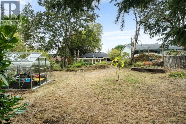 Backyard view with a greenhouse, dry grass, mature trees, and a glimpse of the house. - 14 Eaton Ave, View Royal