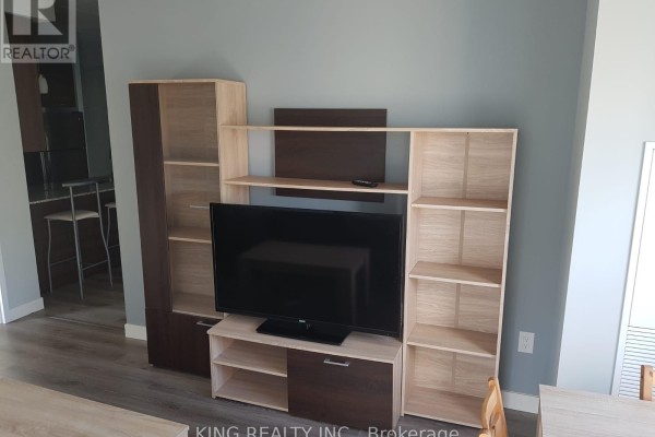 Living area with a large flat-screen TV on a multi-shelf entertainment unit and light wood flooring. - 1405 - 318 Spruce Street, Waterloo