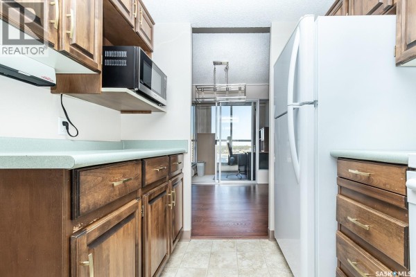 Narrow kitchen view with dark wood cabinets, white refrigerator, sink, and view through doorway to city. - 1405 315 5th Avenue N, Saskatoon