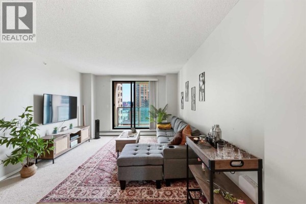 Living room with sectional sofa, coffee table, and large window overlooking city buildings. - 1407, 1111 6 Avenue Sw, Calgary