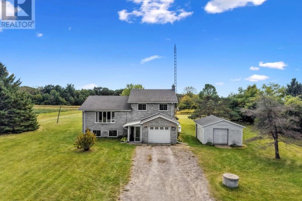 Front view of a two-story grey brick house with a white garage door, set on a wide, cleared lawn. - 141 Cedar Lane, Cramahe