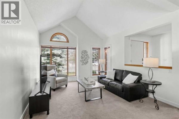 Living room with a vaulted ceiling, black leather sofa, armchair, and large windows overlooking trees. - 14125 Evergreen Street Sw, Calgary
